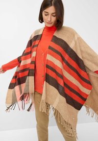 Gestreifter Poncho aus einem Beigemisch aus Wolle in Beige, Rot und Braun. Mit einem fransigen Saum und einem übergroßen Design, über einem orangefarbenen Top getragen.