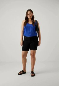 Vrouw in een blauwe mouwloze top, zwarte shorts, zwarte sandalen, met een zwarte schoudertas, staand tegen een effen lichte achtergrond.