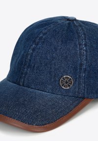 Cappellino in denim blu scuro con visiera in pelle marrone e un emblema sul fronte. Presenta una visiera curva e dettagli cuciti.