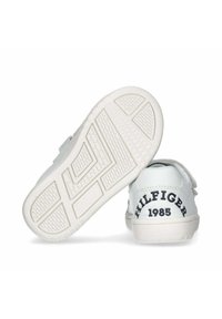 Sneaker bianche con upper in pelle liscia, cinturino in velcro e suola in gomma. Presenta "HILFIGER 1985" in nero sul tallone. Suola testurizzata.