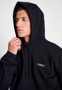 Svart bomullshuvtröja med en framficka, dragsko på huvan och en framträdande logotyp på bröstet. Har ribbade muddarmar och en mjuk textur.