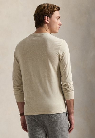 Man with light brown hair wearing a beige long-sleeve shirt and gray textured pants, standing and looking to the side against a neutral background.