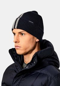 Cappello beanie blu navy con texture a coste e dettaglio grigio, indossato con una giacca imbottita scura, dotata di zip e chiusura a scatto.