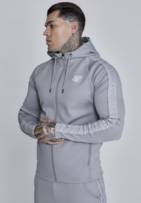 Šedá atletická mikina na zips s jemnou textúrou, ktorá má na hrudi biele logo a vzorované akcenty pozdĺž rukávov.