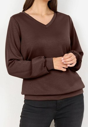 Mujer con blusa marrón de manga larga y escote en V con mangas abullonadas y pantalones negros, con las manos suavemente entrelazadas a la altura de la cintura.