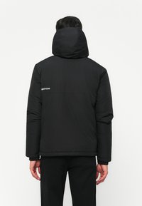 Persoon in een zwarte winterjas met capuchon en zwarte broek, met de rug naar de camera, handen ontspannen naast zich.