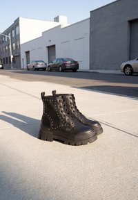 Paire de bottes de combat en cuir noir cloutées avec des semelles épaisses placées sur un trottoir en béton ensoleillé à côté d'une rue où des voitures sont garées.