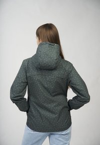 Femme aux longs cheveux bruns portant une veste à capuche vert foncé avec de petits motifs blancs, debout les mains sur les hanches, de dos.