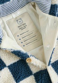 Fleece-Cardigan mit blau-weißem Karo-Muster; ausgestattet mit Holzknöpfen und einem gestreiften Kragen. Pflegeetikett innen mit Namensfeldern.