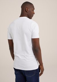 Camiseta blanca de algodón con mangas cortas, cuello redondo y corte recto, llevada con jeans azul oscuro, observada desde la parte de atrás.