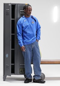 Modrá mikina Nike s dlouhým rukávem a logem, džíny a černé tenisky; stojí vedle šedého šatního skříňky se dvěma otvory.