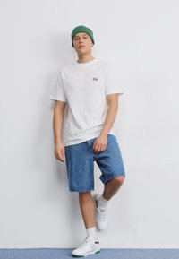 White cotton t-shirt with a small black logo, blue denim shorts, and green knit beanie. White sneakers with green accents.