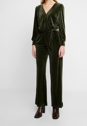 Femme portant une combinaison cache-cœur en velours vert olive à manches longues, avec une ceinture à la taille et un pantalon à jambes larges, associée à des bottines à motifs foncés.