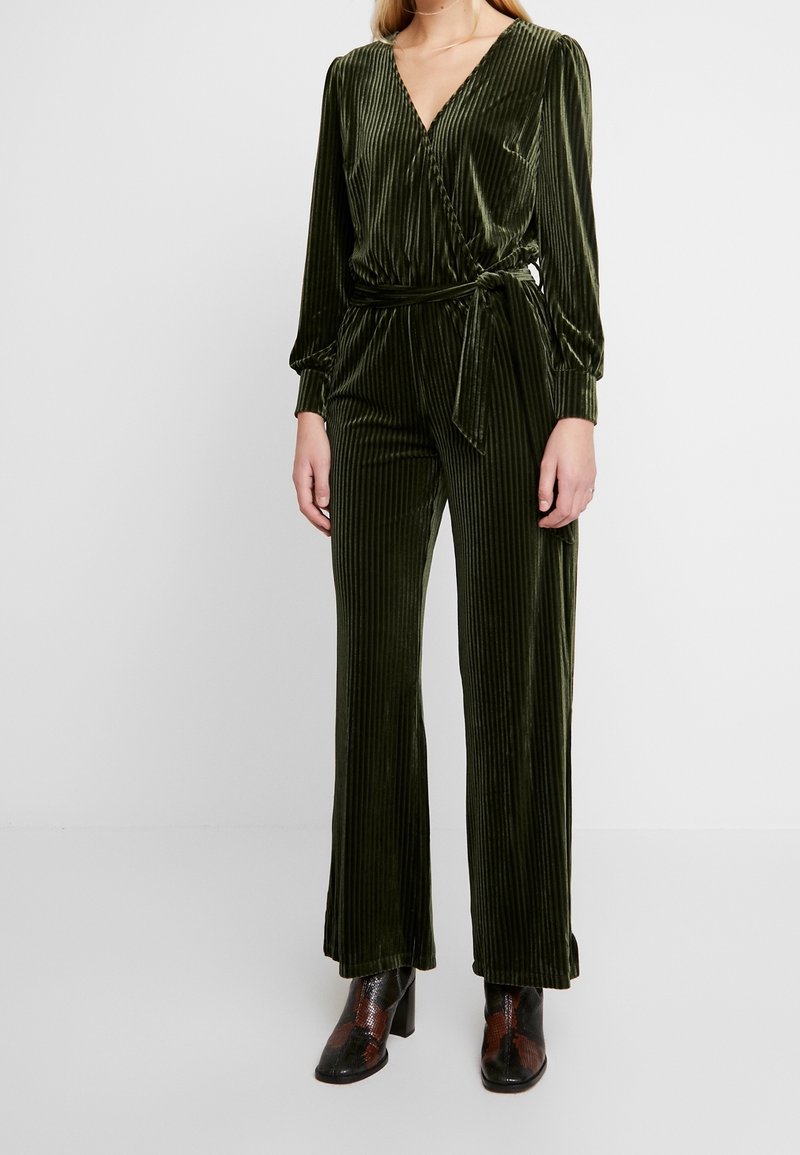 Femme portant une combinaison cache-cœur en velours vert olive à manches longues, avec une ceinture à la taille et un pantalon à jambes larges, associée à des bottines à motifs foncés.