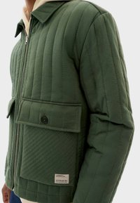 Man in een groen gewatteerd jack met een voorrits en knoopsluiting op de flapzakken, gedragen over een beige hoodie.