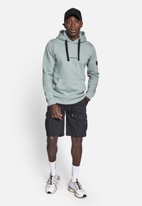 Light teal hoodie with black drawstrings and logo, paired with black cargo shorts. Model wears white socks and athletic shoes with yellow accents.