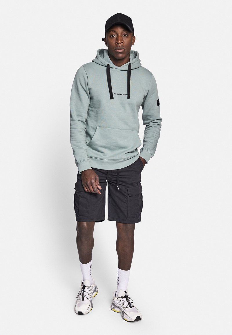 Helles Teal-Hoodie mit schwarzen Kordeln und Logo, kombiniert mit schwarzen Cargo-Shorts. Das Modell trägt weiße Socken und Sportschuhe mit gelben Akzenten.