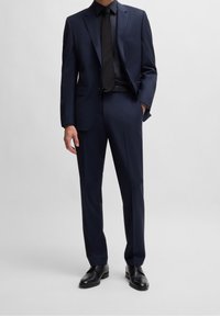 Completo blu navy con design sartoriale, giacca monopetto e pantaloni coordinati. Completato da cravatta nera e scarpe abbinate.