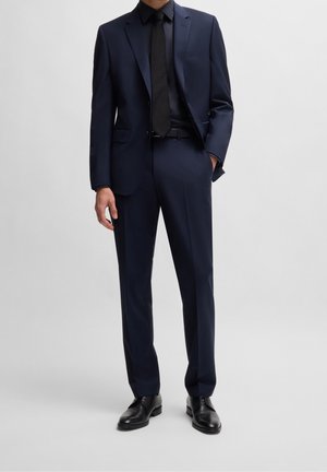 Completo blu navy con design sartoriale, giacca monopetto e pantaloni coordinati. Completato da cravatta nera e scarpe abbinate.