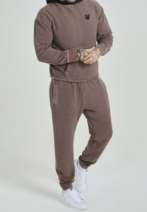 Bruin fleece trainingspak met een capuchon en joggingbroeken met een trekkoord. Bevat een voorzak en geribbelde manchetten. Witte sportschoenen.