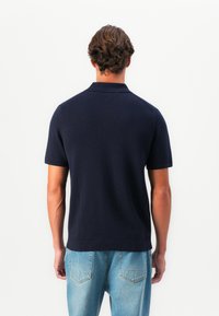 Námorníckom modrá polokošeľa s krátkym rukávom a klasickým golierom, hladká textúra a rovný strih na spodnej strane, nosená s bledomodrými džínsami.