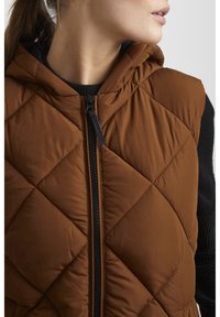 Gilet matelassé marron avec une capuche, doté d'une fermeture éclair noire et de poignets en tricot noir. Matériau lisse avec une texture en motif de diamant.