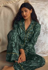 Zelená kvetinová pyžamová sada so žltými lemami, obsahujúca dlhé rukávy a nohavice. Sedenie na pozadí s jemnými, neutrálnymi farbami.