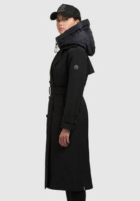 Manteau noir avec un col volumineux noir, une taille ceinturée et des accents de boutons. Le manteau dispose de manches longues et d'une silhouette élégante.