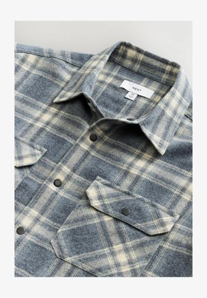 Camisa de franela con un estampado de cuadros en azul y crema, con cuello, dos bolsillos frontales, botones negros y una textura suave. Etiqueta de la marca visible.