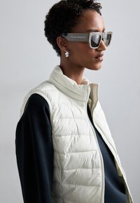 Lagana kremasta pufasta prsluk s visokim ovratnikom, u kombinaciji s crnom dugom majicom. Masivne sive sunčane naočale s oznakom "Alexander McQueen".