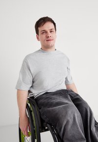 Szara koszulka krótki rękaw i ciemnoszare spodnie na osobie na wózku inwalidzkim. Wózek ma zielone akcenty na kołach.