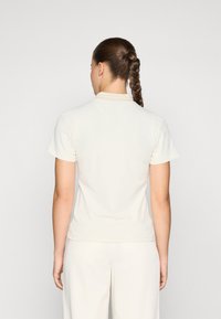 Kremfarget kortermet polo-Shirt laget av et jevnt stoff, med en myk krage og en tettsittende design, vist fra baksiden.