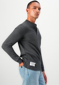 Camasa henley gri cu maneci lungi, textură cu benzi, cu trei nasturi și etichetă laterală. Asortată cu blugi din denim albastru deschis.
