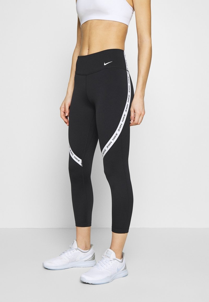 Femme portant un legging noir Nike avec des bandes blanches avec logo sur les côtés, un soutien-gorge de sport blanc et des chaussures de sport blanches, debout sur un sol gris clair.