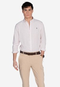 Camisa de botones a rayas rosa clara, hecha de tela con mangas enrolladas, combinada con pantalones cargo beige y un cinturón marrón. Pequeño logo en el pecho.