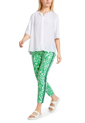 Blouse blanche à col, manches courtes, associée à des leggings verts à motifs avec des bandes latérales noires et des sandales blanches.