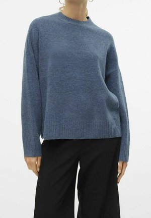 Personne portant un pull en tricot bleu ample à manches longues et un pantalon noir à jambes larges sur fond blanc.