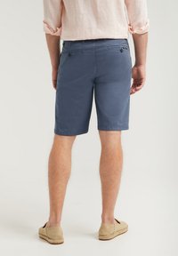 Shorts en coton bleu avec poches arrière, devant plat et bords ourlés, associés à des espadrilles beiges sur un fond neutre.