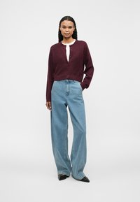 Woman with long curly hair wearing a buttoned burgundy cardigan, white top, wide-leg blue jeans, and black pointed shoes, standing with one hand in pocket.