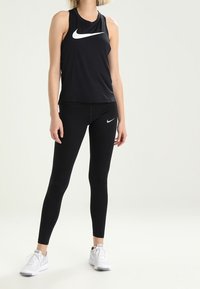 Žena oblečená v černém bezrukávovém tričku Nike a legínách s bílými teniskami Nike na čistém pozadí.