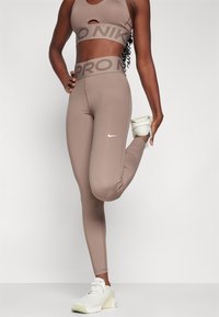 Hnědé, elastické sportovní leginy s vysokým pasem, na kterých je nápis "PRO NIK", v kombinaci s odpovídajícím cropped topem a bílými teniskami.