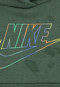 Un hoodie vert foncé présente un logo 'NIKE' réfléchissant avec un swoosh, sur fond de tissu texturé.