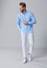 Lichtblauwe button-up shirt met een gladde textuur, gecombineerd met witte slim-fit broek en witte sneakers, tegen een grijze achtergrond.