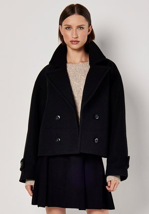 Giubbotto corto doppio petto nero in misto lana, con grandi bottoni e colletto ampio, abbinato a un maglione lavorato a maglia beige.