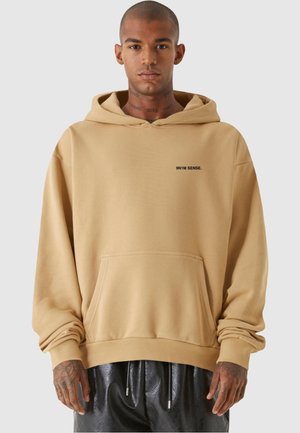 Beige hættetrøje med en lomme foran, der har et lille sort logo på brystet. Stoffet ser blødt ud med en glat tekstur.