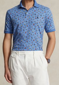 Blå polo skjorta med mönster av segelbåtar i röd, gul och vit, tillsammans med vita veckade byxor och en brun armbandsur.