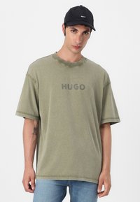 Olive green oversized t-shirt with short sleeves, featuring the text "HUGO" in gray. Worn with light blue jeans and a black cap.