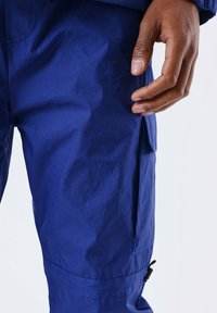 Blauwe nylon broek met een gladde textuur, voorzien van een zijzak en verstelbare enkels. Close-up van de stof en hand.