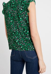 Zelený top s leopardím potiskem, který má volánkové rukávy a volný střih, v kombinaci s tmavě modrými džíny. Materiál vypadá hladce a je lehký.
