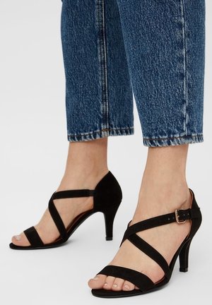 Sorte ruskind højhælede sandaler har krydsende stropper, ankelspænde og et åbent tådesign. Hælen måler cirka 7,5 cm.
